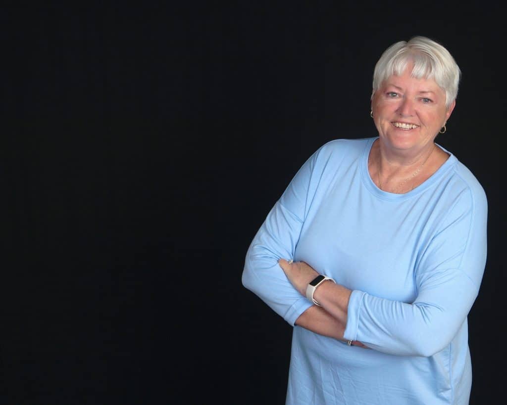 Gail Kenney smiling in blue shirt with arms crossed against dark background